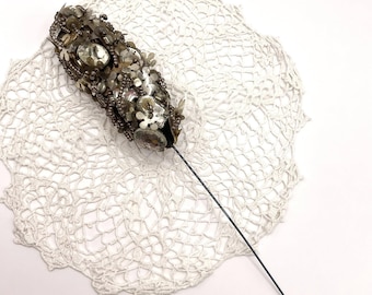 May include: An ornate hat pin with a dark metal shaft. The decorative head is covered in silver and clear beads, sequins, and stones. The pin is set on a white, crocheted doily.