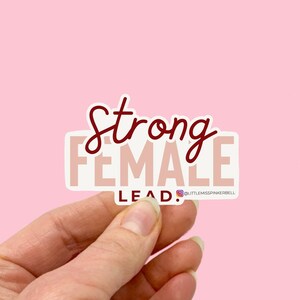 May include: A sticker with the text "Strong Female Lead." The sticker is pink with red lettering and is being held by a hand.