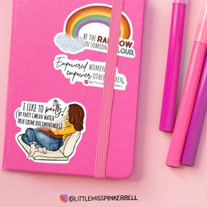 May include: Three stickers on a pink notebook. The top sticker features a rainbow with the text "Be the rainbow in someone's cloud." The middle sticker features the text "Empowered women empower other men." The bottom sticker features a cartoon illustration of a person watching true crime documentaries with the text "I like to party! (By party I mean watch true crime documentaries)".