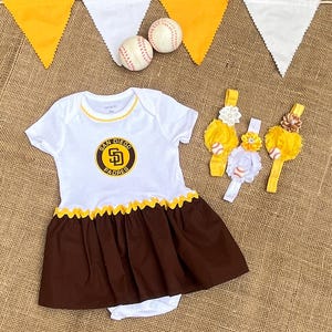 May include: A white and brown baby dress featuring the San Diego Padres logo on the chest, accented with yellow trim. The set includes two yellow and white elastic headbands decorated with fabric flowers and miniature baseballs, plus two baseballs.