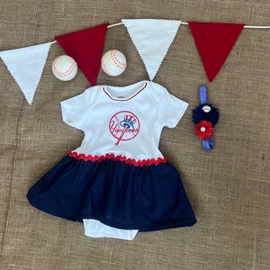 May include: A white and navy blue baby dress with a red scalloped trim, featuring a Yankees logo. Includes a matching headband with a baseball and flowers, and baseballs and pennants. A cute outfit for a baby.