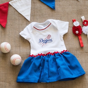 May include: A white and blue baby dress with a Dodgers logo, red trim, and a blue skirt. Two baseballs and two headbands with flower accents are also in the image. A red, white, and blue banner is in the background.