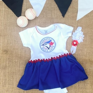 May include: A white baby dress with a Toronto Blue Jays logo, red trim, and a blue skirt. A matching headband with a baseball-themed flower and two baseballs are also present. Black and white pennant banner in the background.
