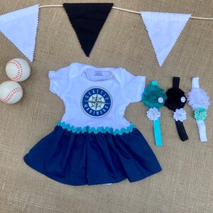 May include: A white and navy blue baby dress with a Seattle Mariners logo, scalloped trim, and a matching headband. The image also includes baseballs and a decorative banner.