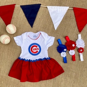 May include: A white and red baby dress with a Chicago Cubs logo, baseballs, and matching headbands. The dress has a red skirt and blue trim. A banner with red, white, and blue pennants is in the background.