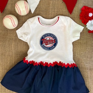 May include: White and navy blue baby dress with a Minnesota Twins baseball team logo. The dress has a red scalloped trim and a navy blue skirt. Two baseballs and red and white pennants are in the background.