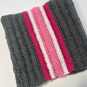 May include: A handmade crochet scarf in a rectangular shape. The scarf is primarily dark gray with a central stripe of pink, white, and fuchsia. The crochet stitch creates a textured appearance, suitable for cold weather.
