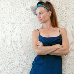 May include: A long, dark teal dress with spaghetti straps and a cowl neckline. The dress is worn by a person with auburn hair and a teal headband. The background is a cream-coloured, crocheted textile.