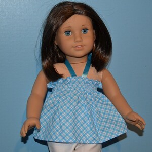 Puede incluir: Una mu&ntilde;eca con un vestido de cuadros azules y blancos con una cinta turquesa. La mu&ntilde;eca tiene el pelo casta&ntilde;o y los ojos azules.