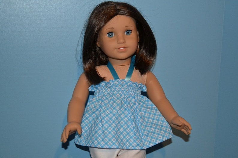 Puede incluir: Una mu&ntilde;eca con un vestido de cuadros azules y blancos con una cinta turquesa. La mu&ntilde;eca tiene el pelo casta&ntilde;o y los ojos azules.