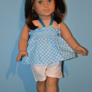 Puede incluir: Una mu&ntilde;eca con cabello casta&ntilde;o que lleva un vestido a cuadros azules y blancos con una correa turquesa y pantalones cortos blancos. La mu&ntilde;eca lleva zapatos turquesa.