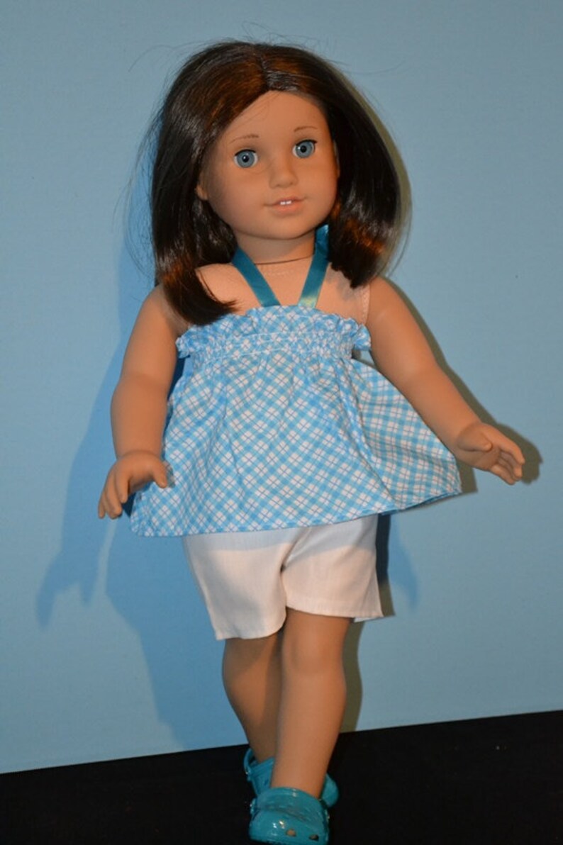 Puede incluir: Una mu&ntilde;eca con cabello casta&ntilde;o que lleva un vestido a cuadros azules y blancos con una correa turquesa y pantalones cortos blancos. La mu&ntilde;eca lleva zapatos turquesa.