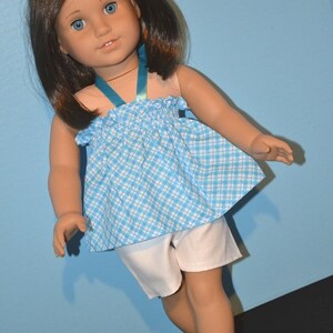 Puede incluir: Una mu&ntilde;eca con un vestido a cuadros azules y blancos con una cinta azul, pantalones cortos blancos y zapatos azules.