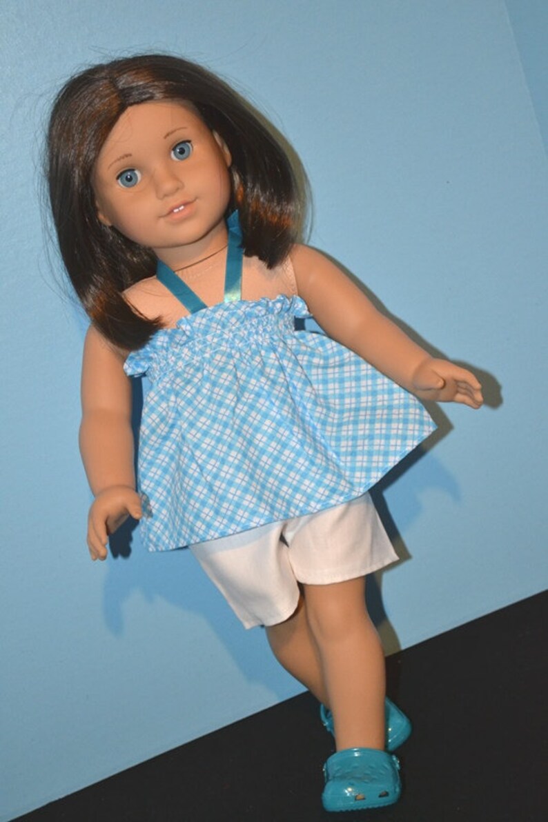 Puede incluir: Una mu&ntilde;eca con un vestido a cuadros azules y blancos con una cinta azul, pantalones cortos blancos y zapatos azules.