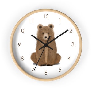 May include: A round wall clock with a light wood frame. The clock face is white with black numerals and hands. A brown bear illustration is centered on the clock face. The bear is sitting upright.