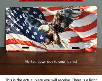 May include: License plate featuring an American flag and a bald eagle in flight. The plate has a red, white, and blue color scheme. The word "CLEARANCE" is at the top. The plate has a small defect.