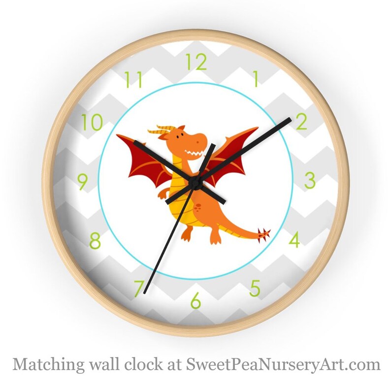 May include: A round wall clock with a light wood frame. The clock face features a cartoon orange dragon with red wings, a blue circle, and a gray and white chevron pattern. Green numbers mark the hours.