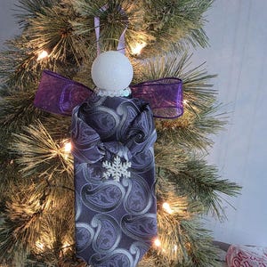Puede incluir: Adorno navideño hecho a mano con forma de ángel. El cuerpo del ángel está hecho de una corbata con estampado de cachemira en tonos morados y grises. Tiene una cabeza blanca brillante, un lazo de cinta morada y un copo de nieve.