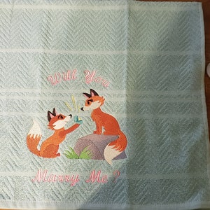 May include: A light blue embroidered towel with a chevron pattern. The towel features two cartoon foxes, one proposing. Text on the towel reads "Will You Marry Me?" in pink script. A romantic gift.
