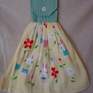 May include: A hanging kitchen towel with a light yellow base, featuring a pattern of cartoon bunnies and flowers in red, white, and blue. The top is a light green fabric with a white button.