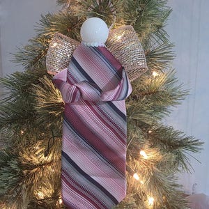 Peut inclure: Ornement de Noël fait à la main en forme d'ange. La tête de l'ange est une boule blanche pailletée, avec un collier de perles. Le corps est fait d'une cravate rayée dans les tons de rose, bordeaux et marine. Les ailes sont en ruban de maille dorée.