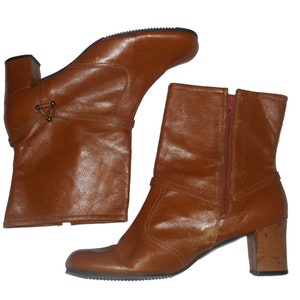 May include: A pair of brown leather ankle boots with a block heel. One boot is upright, and the other is angled. The boots have a side zipper and decorative straps. The heels are approximately 2 inches high.