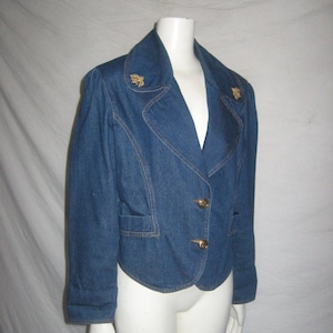 May include: A blue denim jacket with a pointed collar and gold buttons. The jacket has two gold embellishments on the collar.