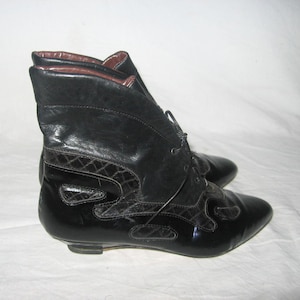 Puede incluir: Botines de cuero negro con punta y cierre de cordones. Las botas tienen un diseño decorativo en el lateral y un interior marrón. Fondo blanco.