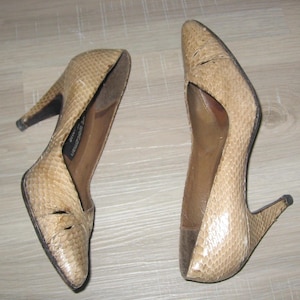 May include: A pair of beige high-heeled shoes with a snakeskin pattern. The shoes have pointed toes and stiletto heels. The heels are approximately 3 inches high. The shoes are on a light brown wooden surface.