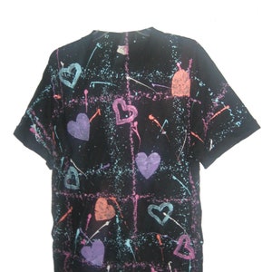 May include: Black t-shirt with a colorful heart and splatter design. The hearts are in shades of purple, orange, and pink, with blue and white paint splatters. The shirt has a grid pattern.