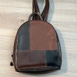 Könnte beinhalten: Ein brauner Lederrucksack mit Patchwork-Design. Der Rucksack hat eine Kombination aus dunkelbraunen, mittelbraunen und schwarzen Lederpaneelen mit weißen Nähten. Er hat einen oberen Griff und verstellbare Schultergurte.