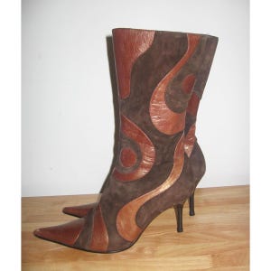 May include: A mid-calf boot featuring a pointed toe and a slim stiletto heel. The exterior is crafted from a combination of dark brown suede and smooth tan leather panels arranged in a swirling, organic pattern that wraps around the entire boot.