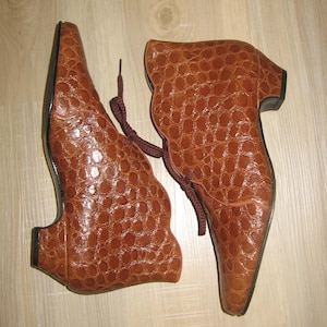 May include: A pair of brown leather ankle boots with a textured, crocodile-like pattern. The boots have pointed toes, short heels, and brown laces. The boots are displayed on a light-colored wooden surface.