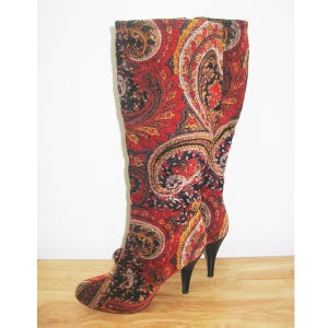 May include: A pair of knee-high boots featuring a vibrant red, black, and gold paisley pattern. These boots are designed with a rounded toe and a slim black stiletto heel. The fabric has a textured appearance with intricate swirling motifs.