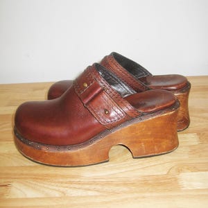 May include: A pair of brown leather clogs with wooden soles. The clogs have a rounded toe, a strap with a buckle, and are adorned with small metal studs. The wooden platform sole adds height.