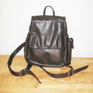 May include: A dark brown leather backpack with a flap closure, adjustable straps, and a top handle. The backpack has a small side pocket and silver-toned hardware. The bag is sitting on a wooden surface.