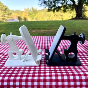 May include: A white and a black phone stand, each shaped like a vintage sewing machine. The stands feature a flat rectangular base, a support arm for a mobile device, and a decorative handwheel on the side, designed to hold a phone upright.