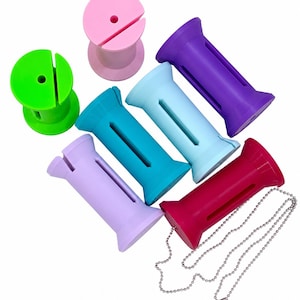 May include: A collection of seven cylindrical plastic spools in various colors including pink, lime green, purple, light blue, teal, lavender, and magenta. Each spool features a vertical slit and a central hole, with one attached to a silver ball chain.