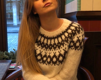 May include: A woman wearing a white sweater with a brown and white geometric pattern. The sweater has a round neckline and long sleeves. She is sitting on a wooden chair with her arms crossed.