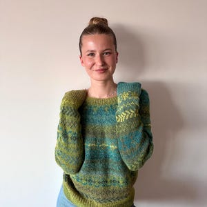 May include: A woman wearing a green and blue patterned jumper with a high neckline and long sleeves. She is wearing blue jeans and has her hair pulled back in a bun.