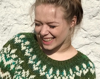 May include: A woman with brown hair in a bun is wearing a green, white, and blue knitted sweater with a traditional Scandinavian pattern. She is smiling and looking down.