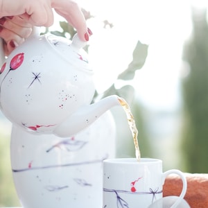 May include: A white teapot with a floral design is pouring tea into a white mug with a floral design. The teapot and mug are on a white table with a loaf of bread in the background.