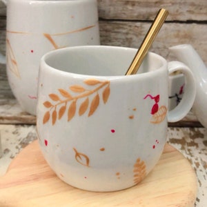 Peut inclure: Tasse en céramique blanche avec une cuillère dorée. La tasse présente des motifs de feuilles dorées et des éclaboussures rouges. Une tasse assortie et un petit pichet sont visibles en arrière-plan. La tasse est posée sur un dessous de verre en bois.