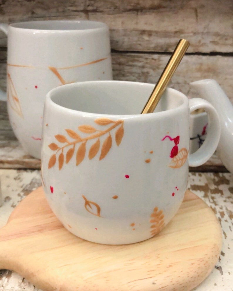 May include: White ceramic mug with a gold-colored spoon inside. The mug features gold leaf and red splatter designs. A matching mug and a small pitcher are visible in the background. The mug is sitting on a wooden coaster.