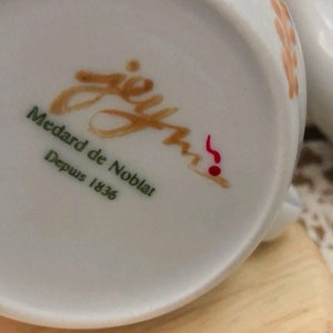 May include: Close-up of a white porcelain teacup base with the text "jey Medard de Noblat Depuis 1836" in brown and green. A red musical note is also visible. The cup has a light brown floral design.
