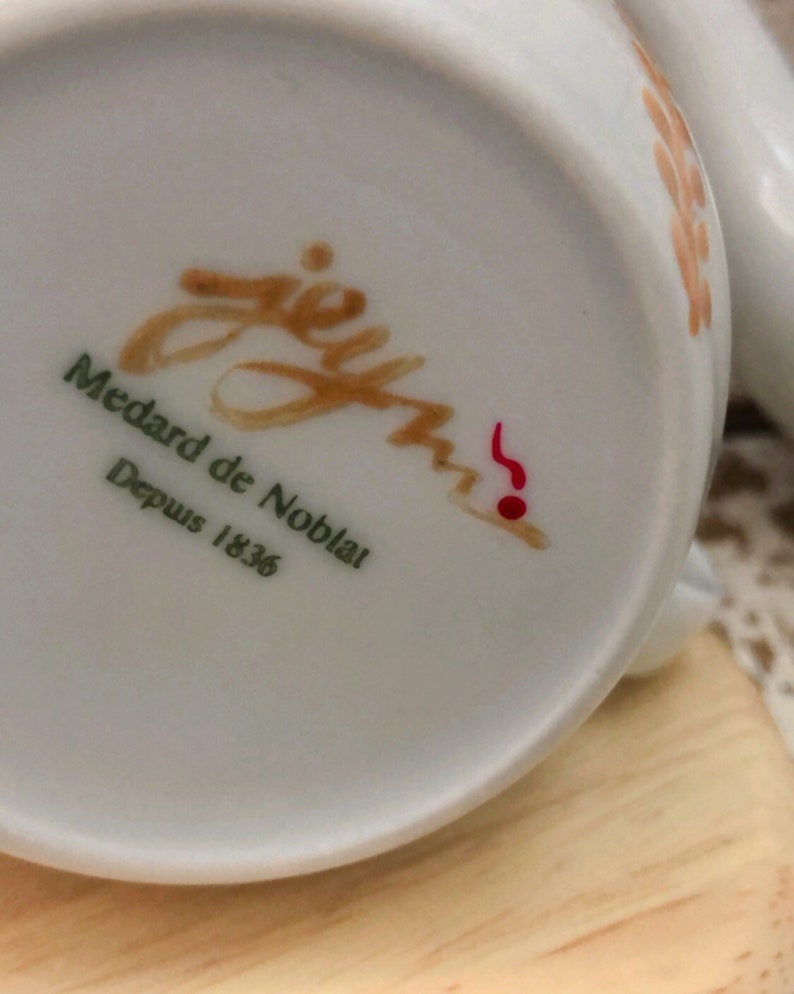 May include: Close-up of a white porcelain teacup base with the text "jey Medard de Noblat Depuis 1836" in brown and green. A red musical note is also visible. The cup has a light brown floral design.
