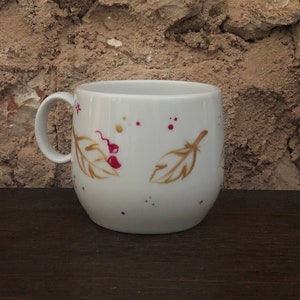 May include: White ceramic mug with a curved body and a handle. The mug is decorated with gold-colored feather and berry designs, along with red and gold paint splatters. The mug sits on a dark brown wooden surface.