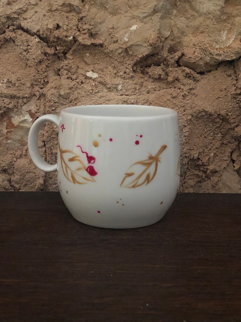 May include: White ceramic mug with a curved body and a handle. The mug is decorated with gold-colored feather and berry designs, along with red and gold paint splatters. The mug sits on a dark brown wooden surface.
