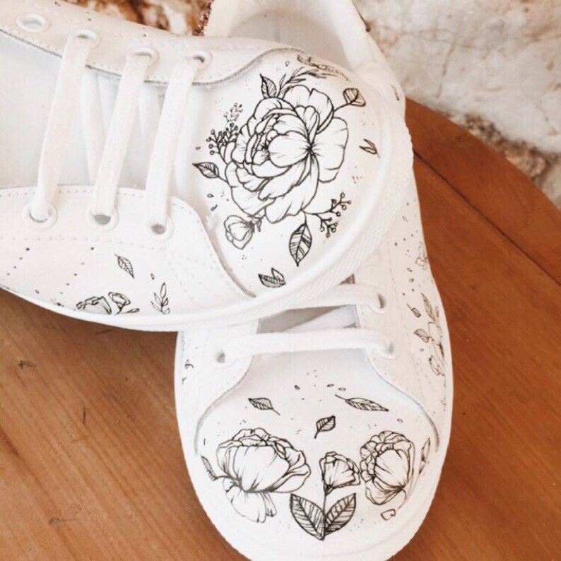 May include: A pair of white canvas trainers with black floral designs painted on them. The flowers are stylised and include peonies and leaves.