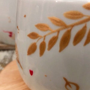 May include: Close-up of a white ceramic mug with a gold leaf design. The mug has a rounded shape and is decorated with gold-colored leaves and small red heart accents. The mug is on a wooden surface.